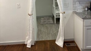 A room with a wooden floor, white walls, a kitchen area with a granite countertop, and a doorway with sheer curtains. - Modern Beauty in Crestline Park (Birmingham)