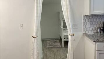 A room with a wooden floor, white walls, a kitchen area with a granite countertop, and a doorway with sheer curtains.
