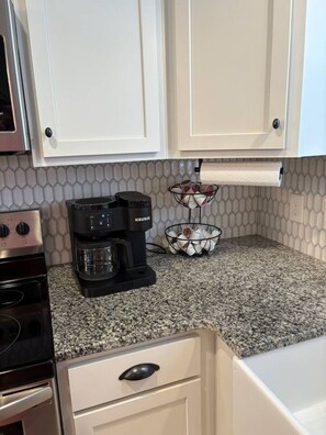 Une cuisine avec des meubles blancs, une machine à café Keurig, un panier de capsules de café, une cuisinière en acier inoxydable et un comptoir en granit.