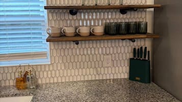 A kitchen with a granite countertop, a dishwasher, and a set of shelves with glassware and kitchen utensils.
