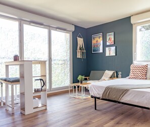Un dormitorio con cama, escritorio y silla. Hay dos ventanas con cortinas, un estante en la pared y un tapiz de macramé.