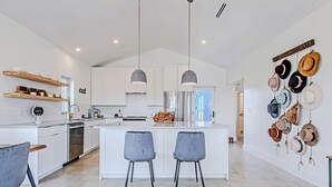 A modern kitchen with white cabinetry, a central island with bar stools, pendant lighting, and a wall-mounted hat display.