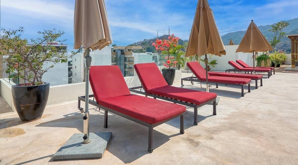 A rooftop lounge area with sun loungers and umbrellas, potted plants, and a view of buildings and hills.