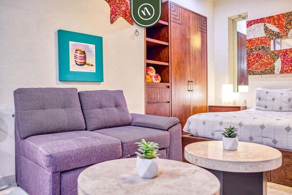 A modern room with a grey sofa, a bed with patterned bedding, a small round table with a plant, and a wooden cabinet with shelves.