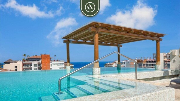 Un espace piscine agrémenté d’une pergola en bois, d’une eau d’un bleu limpide et offrant une vue sur l’océan et les bâtiments qui se dressent à l’arrière-plan.
