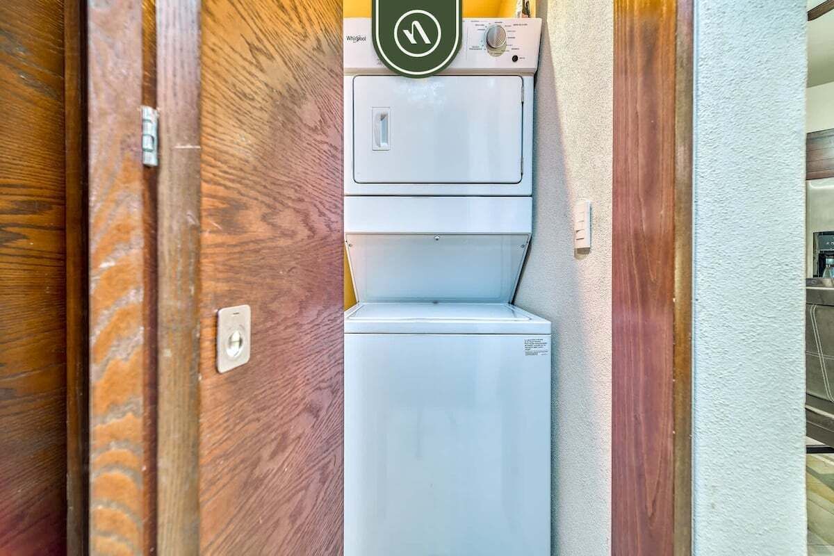 Una unidad compacta de lavadora y secadora, blanca y fijada a la pared, en un estrecho pasillo con puertas de madera y una pared blanca.
