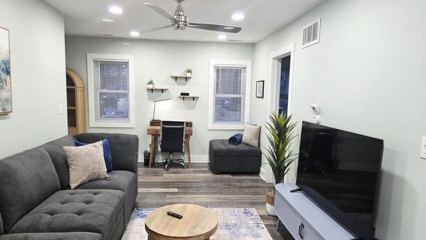 A modern living room with a grey sofa, a round wooden coffee table, a flat-screen TV on a stand, and a ceiling fan. - Charlie`s by the Bay #3 (New Baltimore)