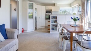 Ein kompakter Wohnraum mit Küchenzeile, einem Essbereich mit Holztisch und Stühlen, einer Couch mit blauem Kissen und einem kleinen Fenster mit Vorhängen.