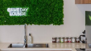 Eine Küche mit Spüle aus Edelstahl, Holdekor-Schränken, einer grünen Wand mit dem Schriftzug 'GAME DAY LOUNGE' und verschiedenen Gewürzgläsern auf der Arbeitsfläche.