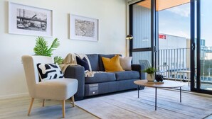 A modern living room with a sofa, armchair, and coffee table, featuring artwork on the walls and a view of the city through large windows.