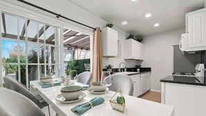 A modern kitchen with white cabinets, a dining area with a white table set for four, and a large window with a view of greenery. - PB Hot Tub Triplex 5BR, MiniGolf, Games & Parking! (San Diego)
