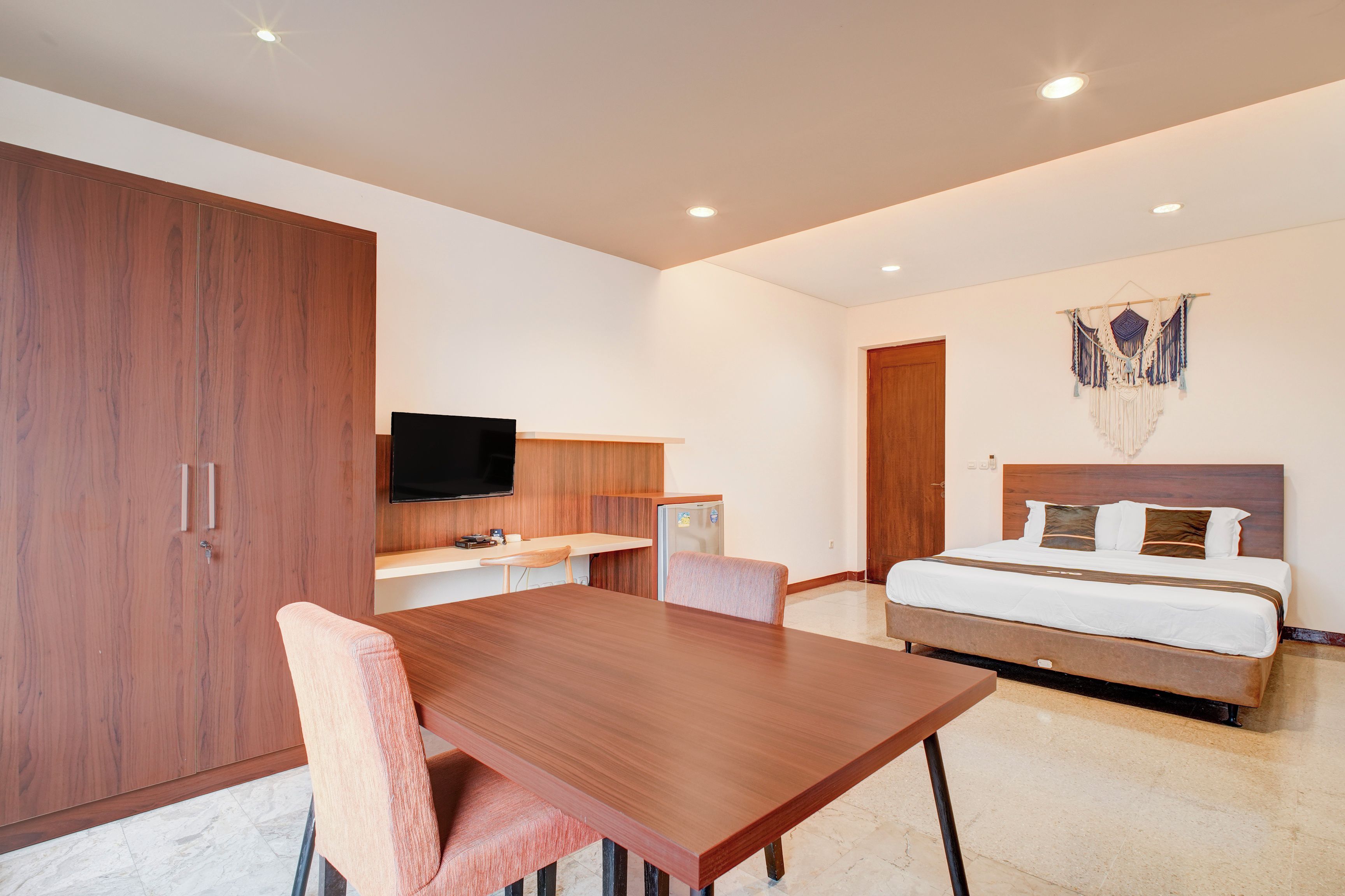 Quarto de hotel moderno com uma mesa de jantar de madeira, uma cama com cabeceira, uma TV de tela plana na parede e um detalhe decorativo em macramé sobre a cama.