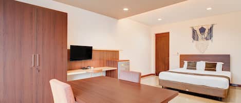 A modern hotel room with a wooden dining table, a bed with a headboard, a flat-screen TV mounted on the wall, and a decorative macrame piece above the bed.