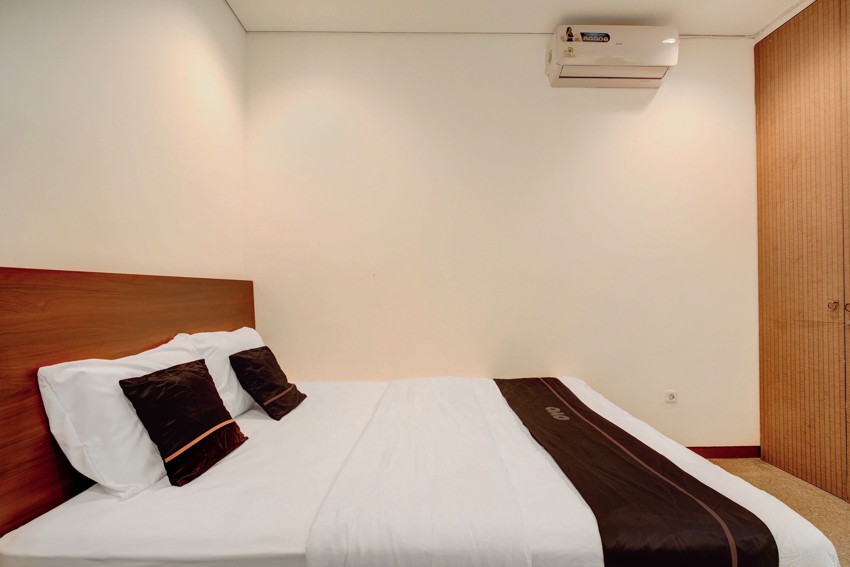 A hotel room with a neatly made bed, a wooden headboard, a wall-mounted air conditioner, and a wooden wardrobe.