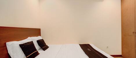 A hotel room with a neatly made bed, a wooden headboard, a wall-mounted air conditioner, and a wooden wardrobe.