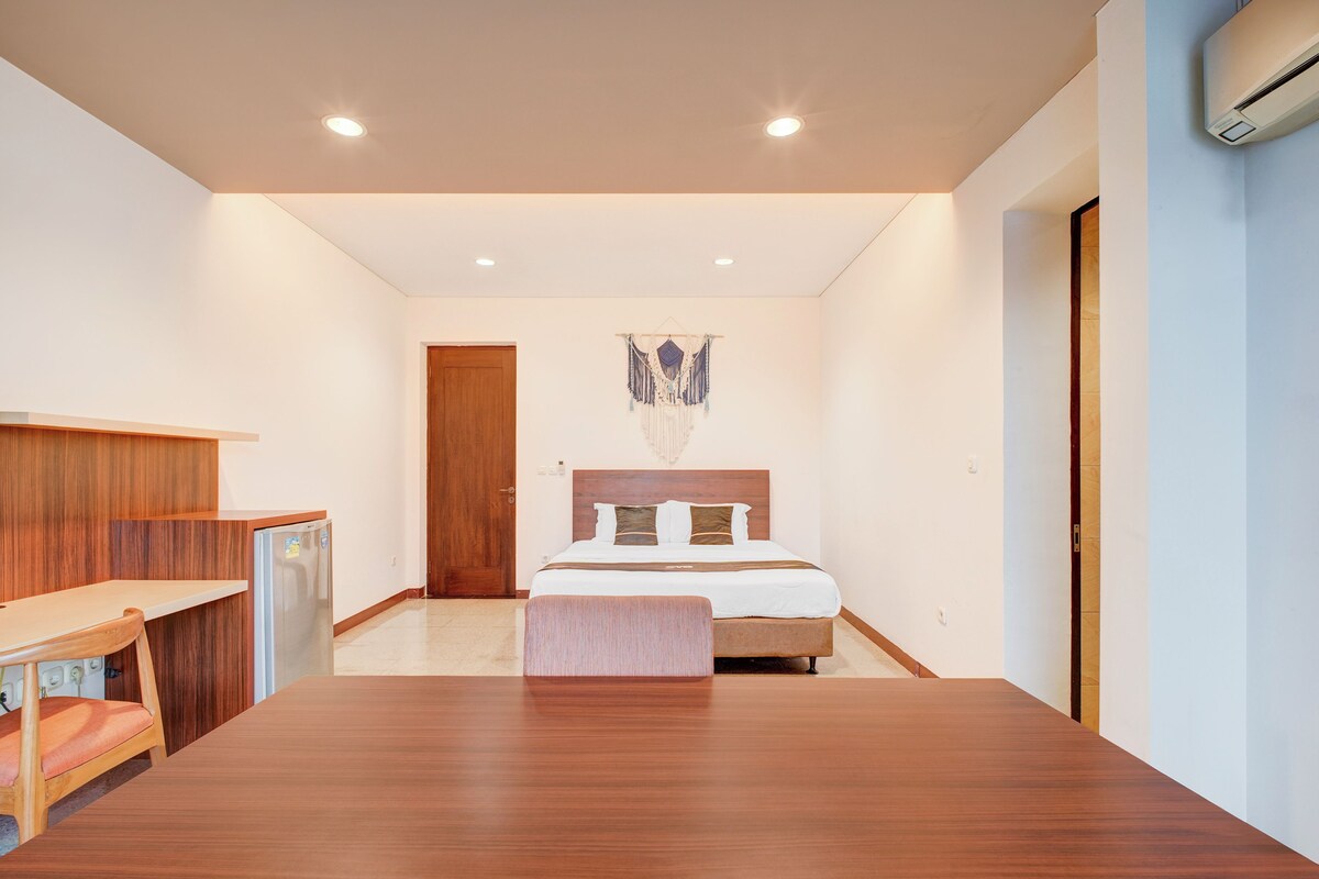 A modern hotel room with a wooden desk, a bed with a headboard, a wall-mounted macrame decoration, and a built-in air conditioning unit.