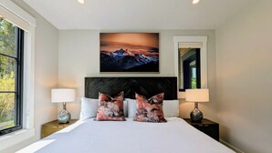 Une chambre à coucher avec un grand lit, deux coussins décoratifs, une table de chevet avec une lampe, un tableau encadré au mur et une fenêtre donnant sur des arbres.