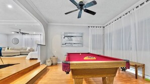 A pool table with a red felt surface, a ceiling fan, and a light fixture in a room with wooden flooring and a white couch. - Massive 5BR Resort Style Retreat with Heated Pool (San Diego)