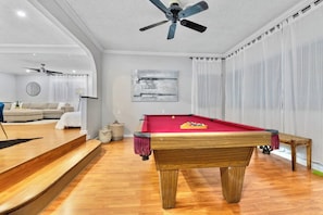 Ett biljardbord med en röd filtduk, en takfläkt och en lampa i ett rum med trägolv och en vit soffa.