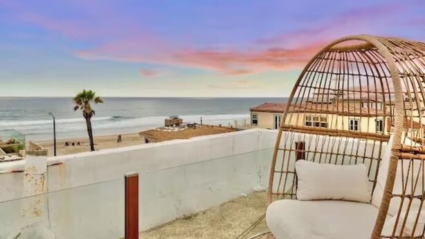 A balcony with a wicker chair overlooking a beach and a building.