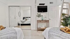 A bedroom with two beds, a TV mounted on the wall, a sliding door leading to another room, and a small table with a vase and a plant.