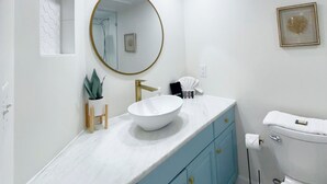 A bathroom with a white sink, blue cabinets, a gold faucet, a round mirror, and a toilet.