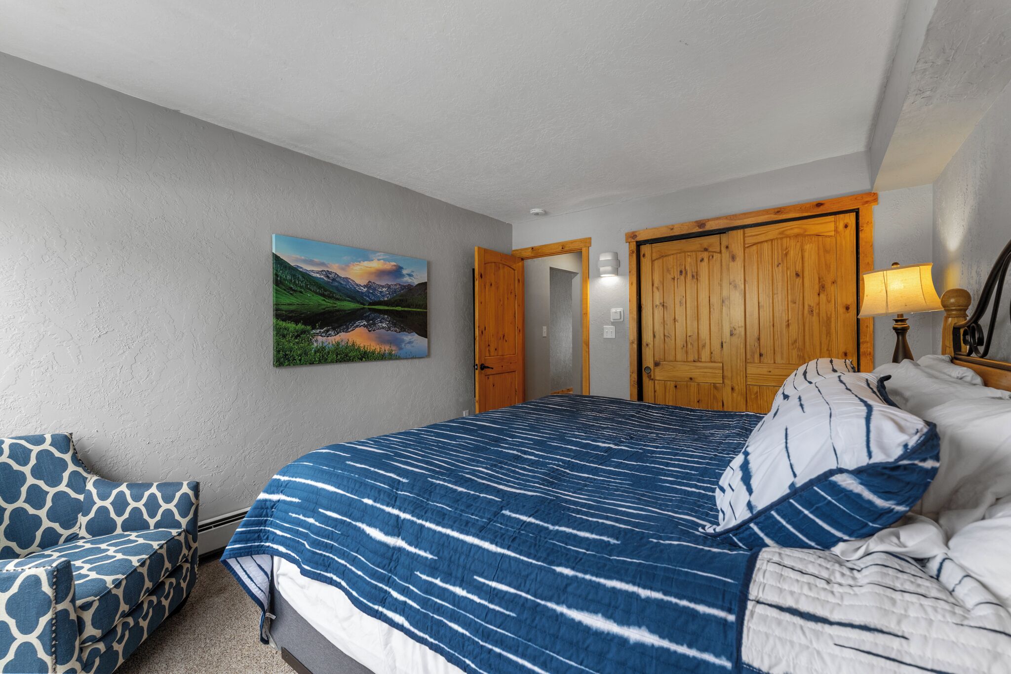 A bedroom with a large bed, a chair, a wooden wardrobe, and a painting on the wall.