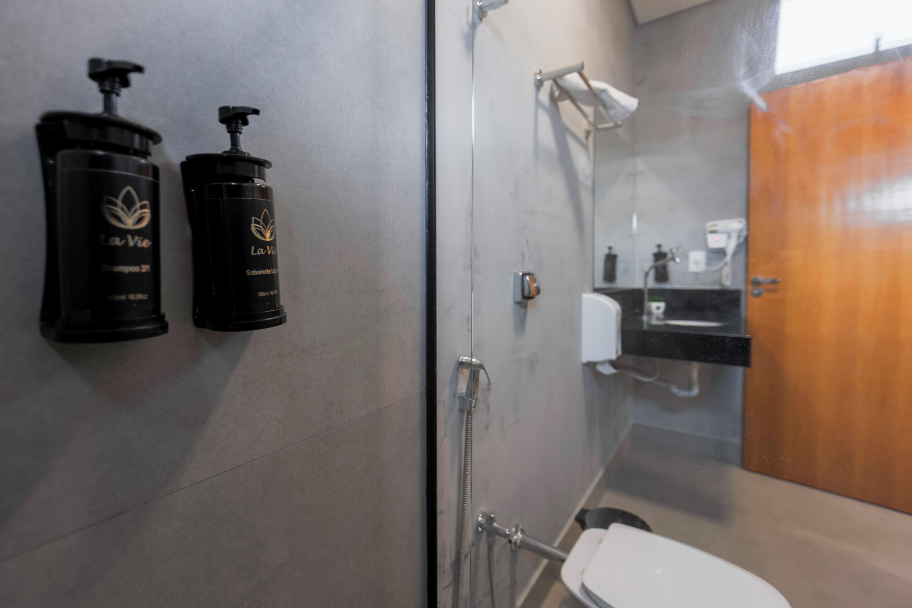 Un baño con cabina de ducha de vidrio, un inodoro blanco y dos botellas de champú La Vie negras fijadas a la pared.