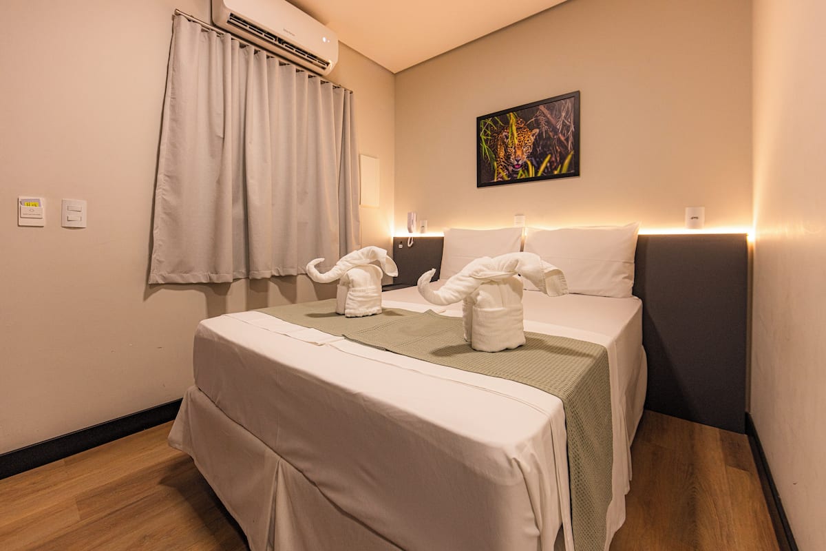 A hotel room with two beds, a wall-mounted air conditioner, a curtain, a painting, and a bedside table with a lamp.