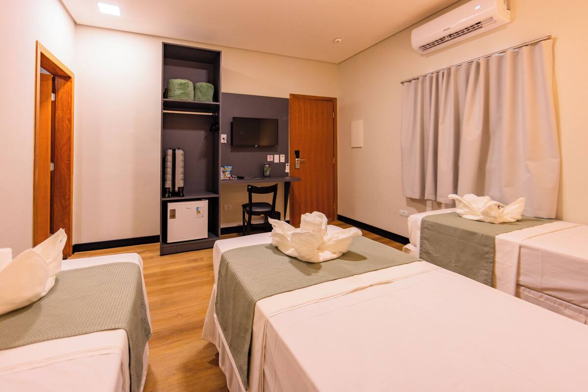 A hotel room with two single beds, a desk with a chair, a television, a wardrobe, and an air conditioning unit.
