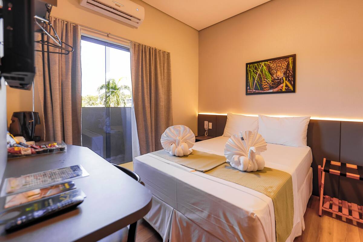 A hotel room with a bed, a desk with a chair, a TV, and a window with curtains.