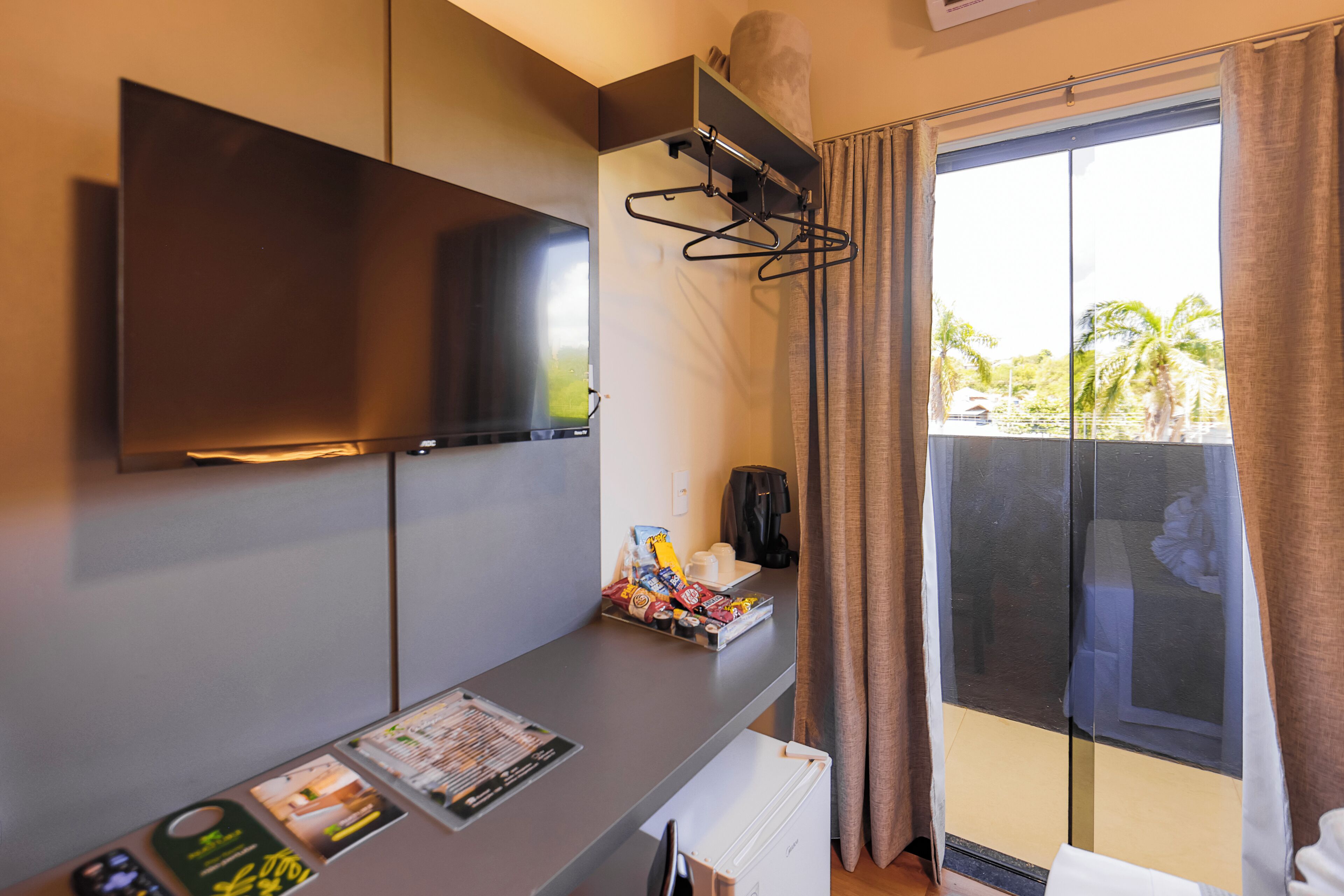 A hotel room with a flat-screen TV mounted on the wall, a coffee maker, a tray with snacks, and a view of palm trees through the window.