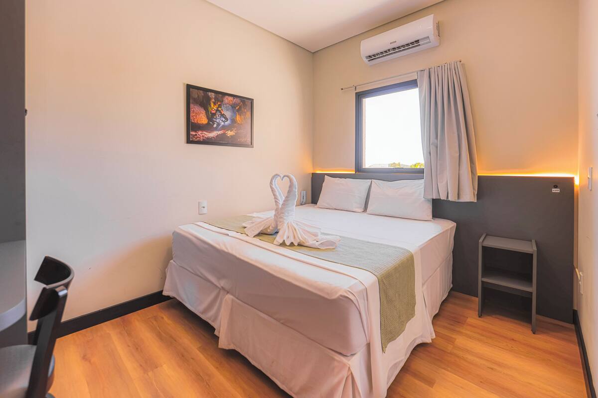 A hotel room with a bed, a window with curtains, an air conditioning unit, a small table, and a chair.
