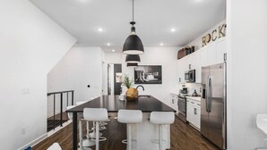 Una cocina moderna con una isla central, taburetes blancos de bar, electrodomésticos de acero inoxidable y una lámpara colgante negra.