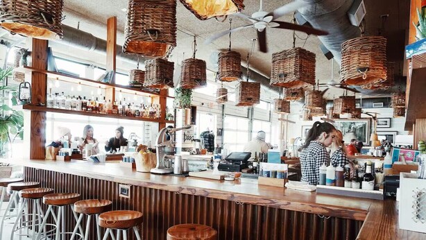 Un bar con taburetes de madera, una barra con caja registradora y cestas colgantes.