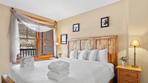 Un dormitorio con una cama grande, cabecera de madera, mesita de noche con lámpara y ventana con cortinas.