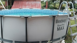 Een stalen Pro MAX bovengronds zwembad met een blauw mozaïek betegelde binnenkant, omringd door een tuin met een schommelset en een gebouw met een rood dak.