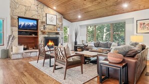 A cozy living room with a stone fireplace, wooden ceiling, and a large window offering a view of trees. - Family-Friendly 5BR | Gather, Relax & Explore (Park City)