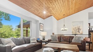 A modern living room with a wooden ceiling, large windows, a brown leather sofa, a wooden coffee table, and a view of greenery outside. - Family-Friendly 5BR | Gather, Relax & Explore (Park City)