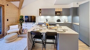 A modern kitchen with a central island, built-in appliances, and a dining area with a wicker chair and ottoman. - Tauari - Les Gets - Les Perrières (Les Gets)