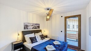 A bedroom with a bed, a nightstand, a wall-mounted lamp, a painting, and a door leading to an outdoor area. - Tauari - Les Gets - Les Perrières (Les Gets)