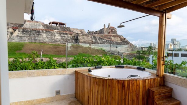 A wooden hot tub on a rooftop terrace with a view of a rocky hill and city buildings. - Blue House Castillo View – Private Jacuzzi & Colonial Balconies (Cartagena)
