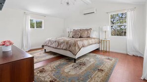 Un dormitorio con una cama grande, una araña, aire acondicionado, una ventana con cortinas y una pequeña mesa con lámpara.