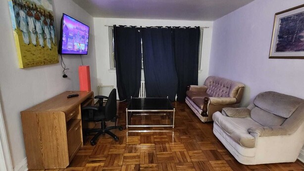 Una sala de estar con un escritorio de madera, una silla de oficina negra, una mesa de centro de vidrio, un sofá color beige y un televisor montado en la pared.
