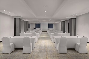 Ein Konferenzraum mit Reihen aus weißen Tischen und Stühlen, einer Projektionsleinwand und einem Fliesenboden.