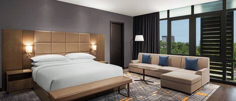 A modern hotel room with a large bed, a sofa, a bench, a nightstand, and a lamp.