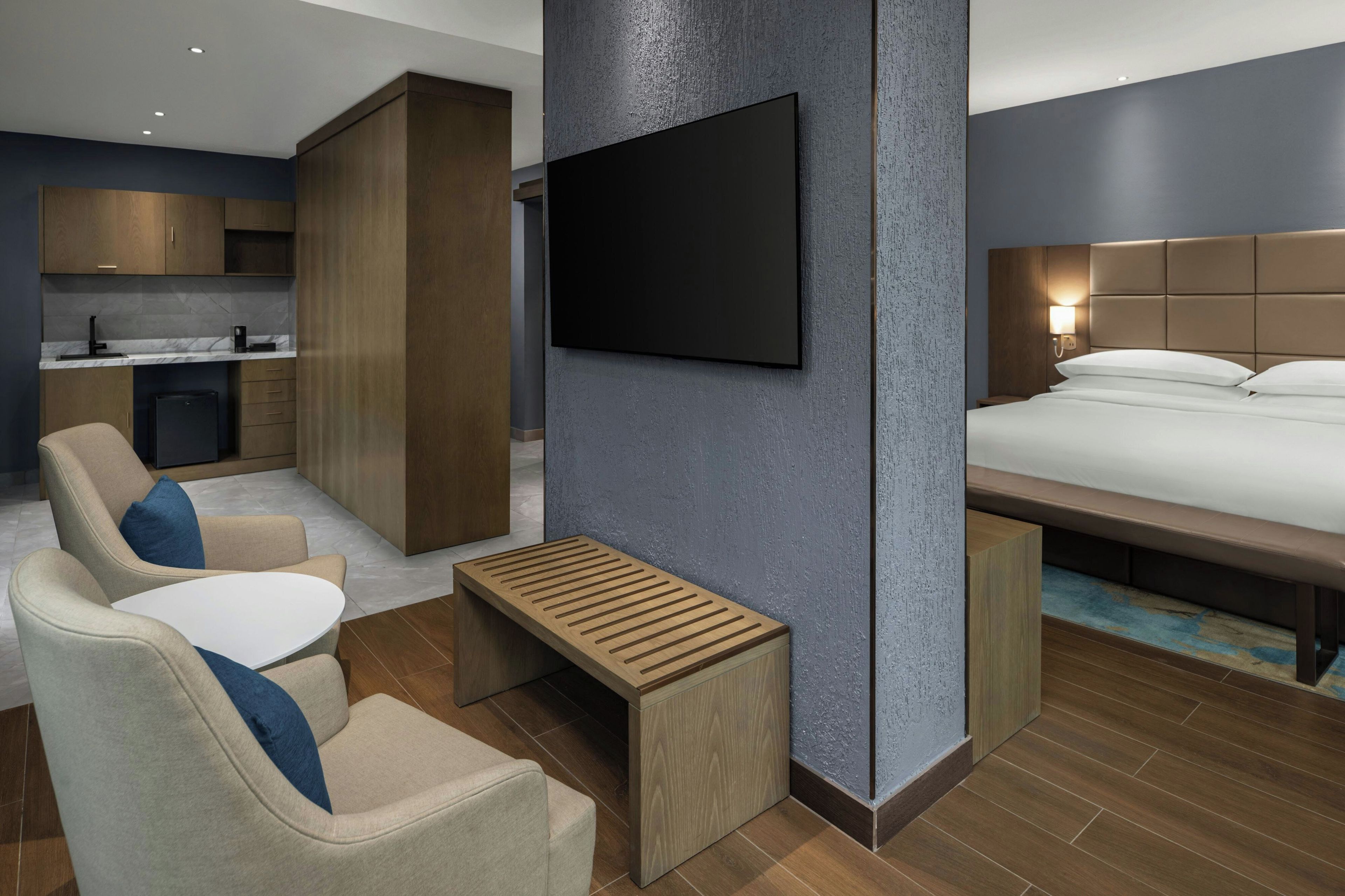 Una habitación de hotel moderna con una cama, un televisor montado en la pared, un espacio de descanso con una mesa y una kitchenette.