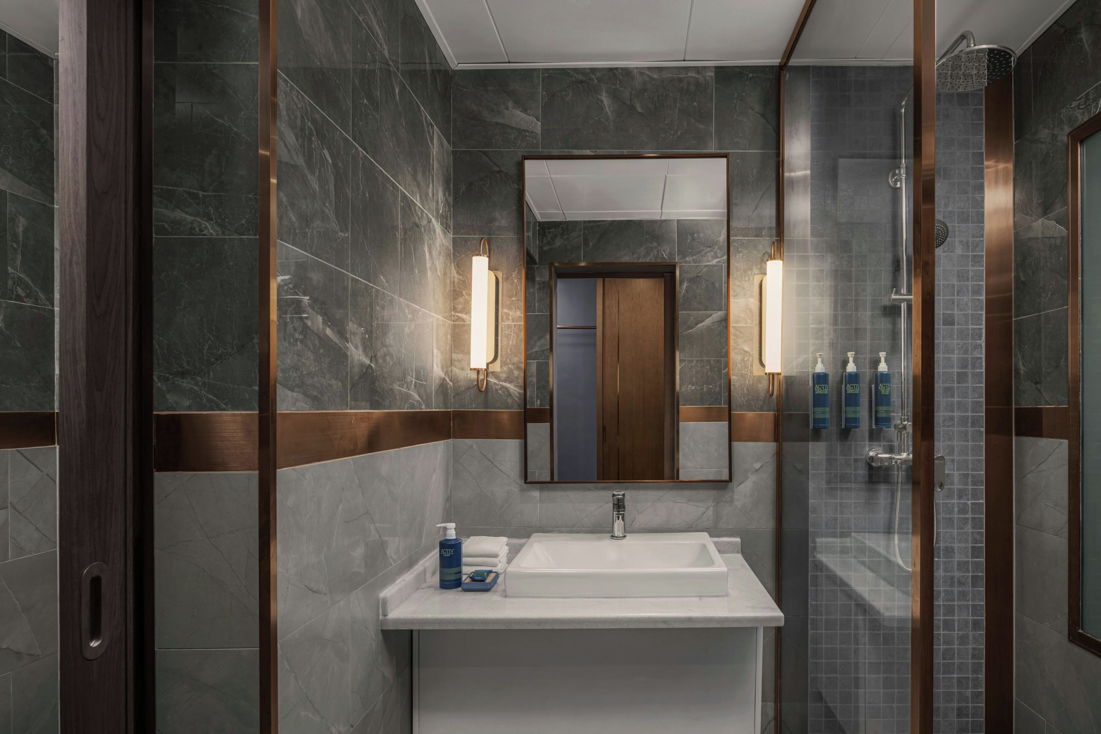 A modern bathroom with a marble wall, a large mirror, a white sink, and a glass-enclosed shower area.