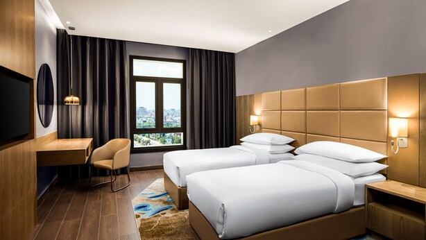 A hotel room with a large bed, a desk, a chair, a TV, and a window with a city view.