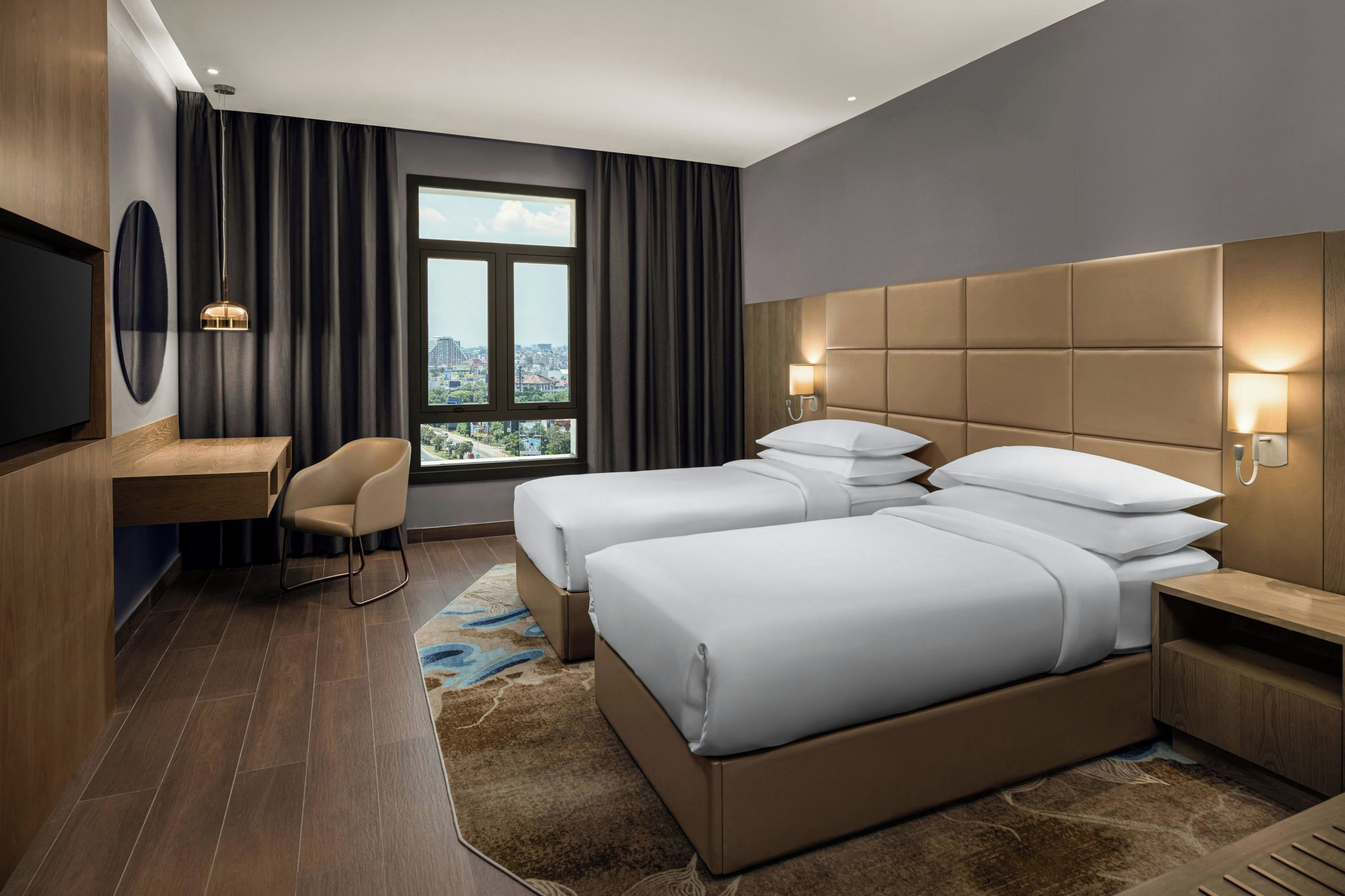 A hotel room with a large bed, a desk, a chair, and a view of the city through a window.